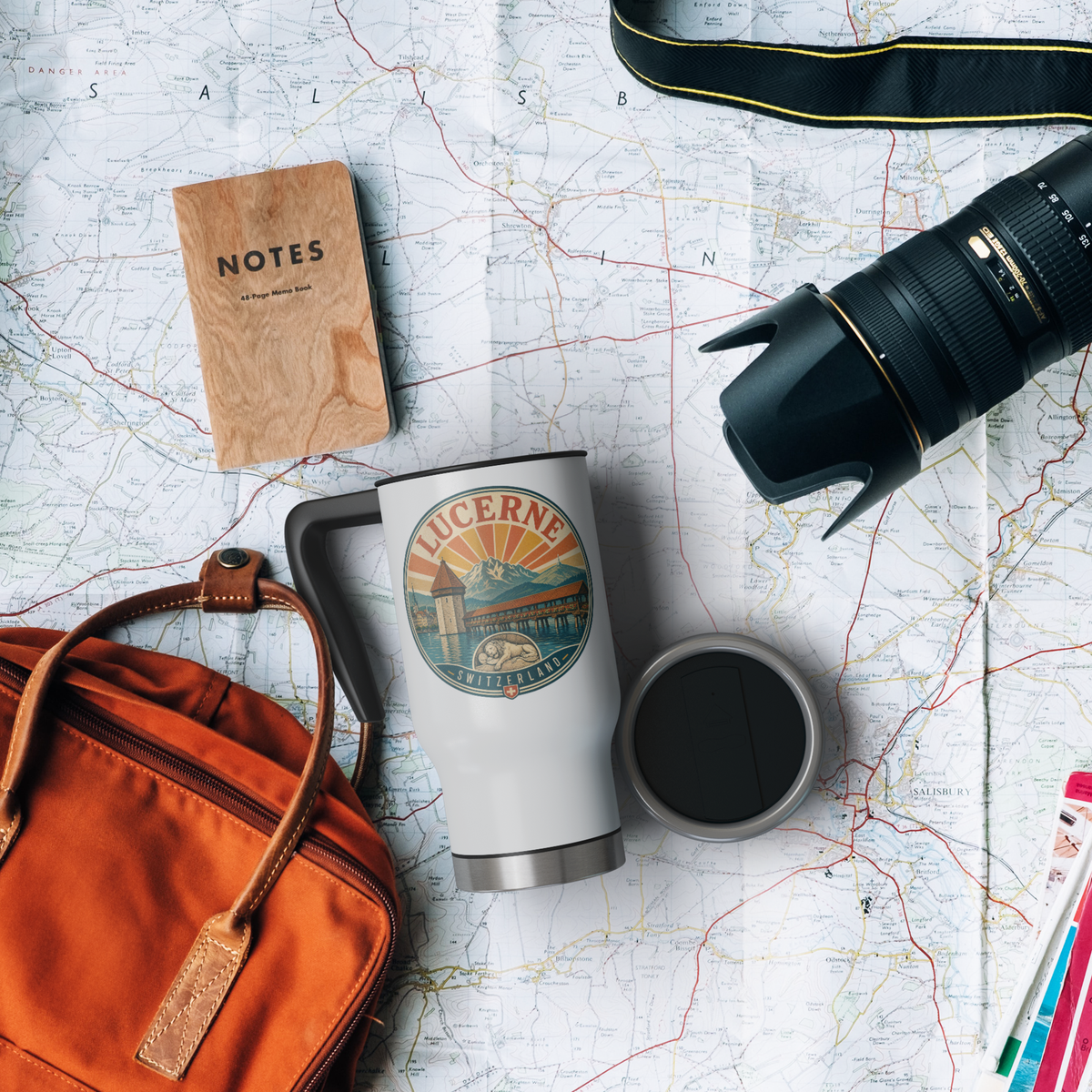Overhead view of a Lucerne Switzerland travel mug and lid on a map beside a camera lens, notebook, and travel bag.