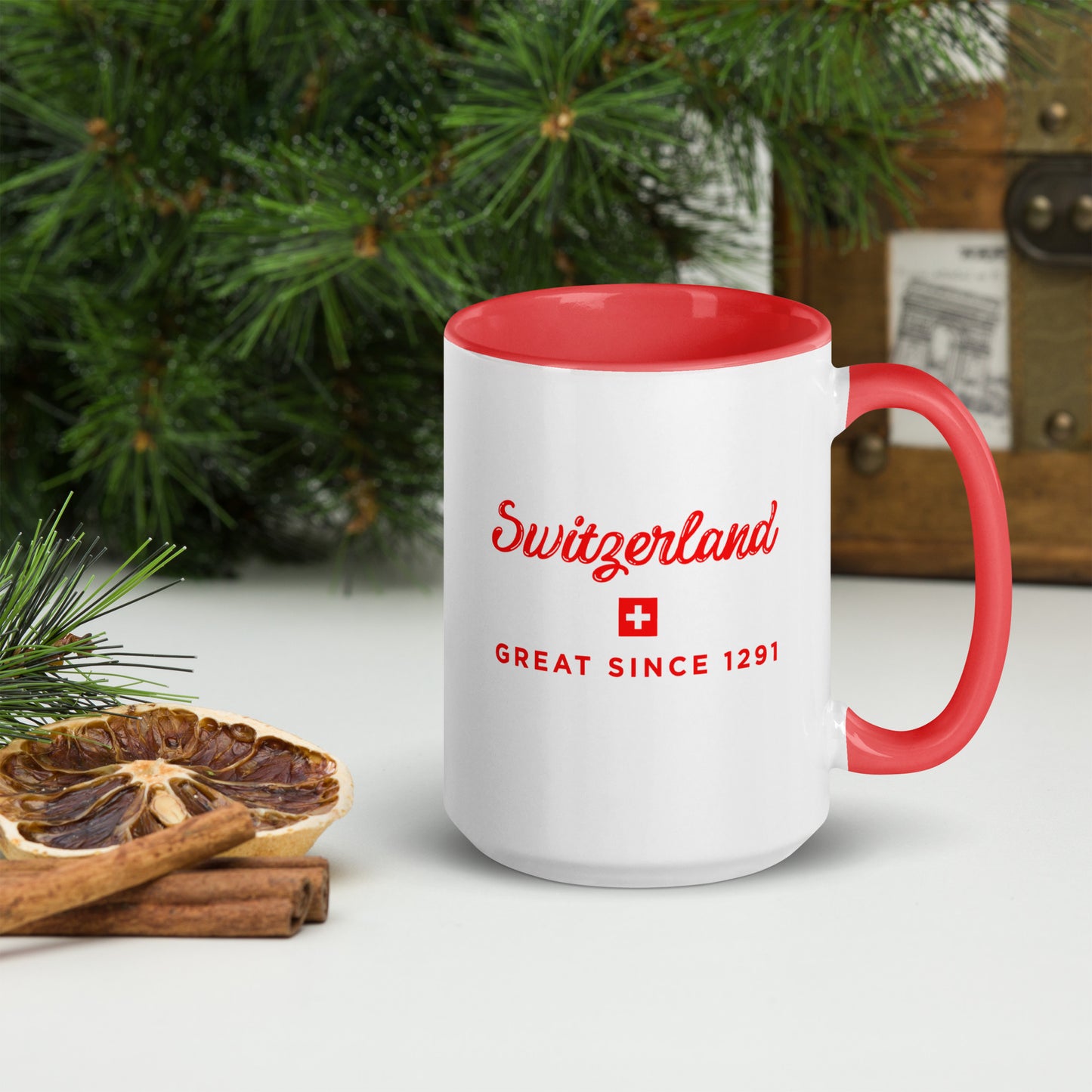 A white ceramic mug with a red interior, red handle, and red printed text reading 'Switzerland' and 'GREAT SINCE 1291' with a small Swiss flag, styled on a tabletop with pine branches and a dried orange slice.