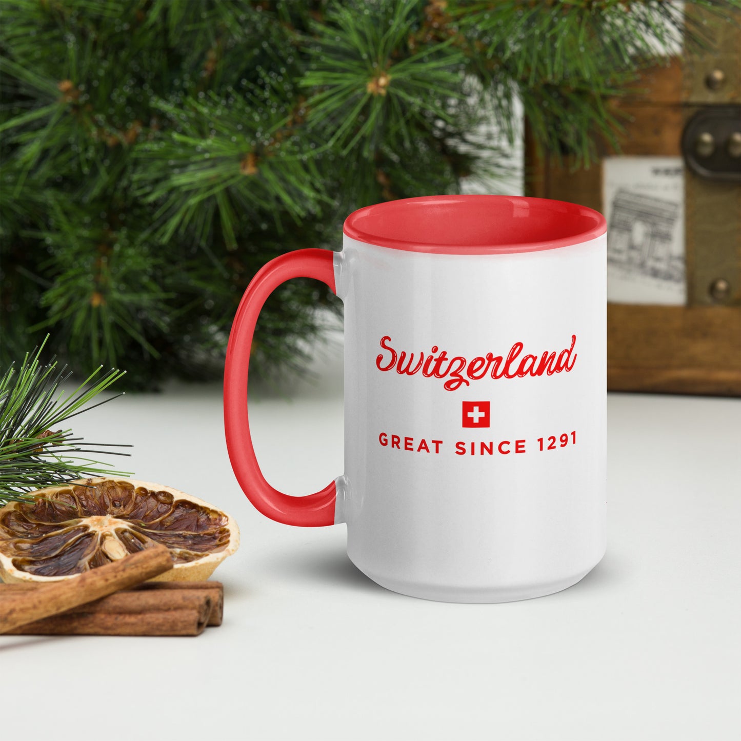 A white ceramic mug with a red handle and red interior, printed with 'Switzerland' and 'GREAT SINCE 1291' in red text alongside a small Swiss flag. The mug is styled on a tabletop with a dried orange slice, cinnamon sticks, green pine branches, and a rustic wooden box in the background.
