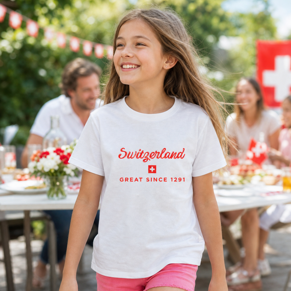 A young girl smiling outdoors while wearing a white t-shirt with 'Switzerland GREAT SINCE 1291' printed in red, set against a blurred background of a garden party.