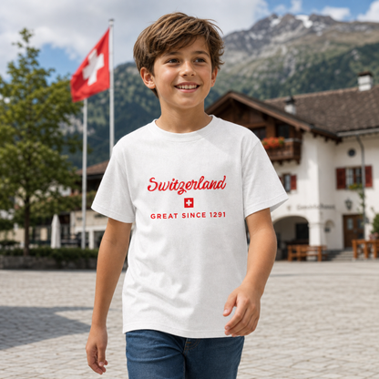 A young boy smiling in an outdoor alpine town square, wearing a white t-shirt with a red 'Switzerland GREAT SINCE 1291' graphic on the chest. Switzerland Great Since 1291 kids t-shirt – lifestyle mockup, casual outdoor wear