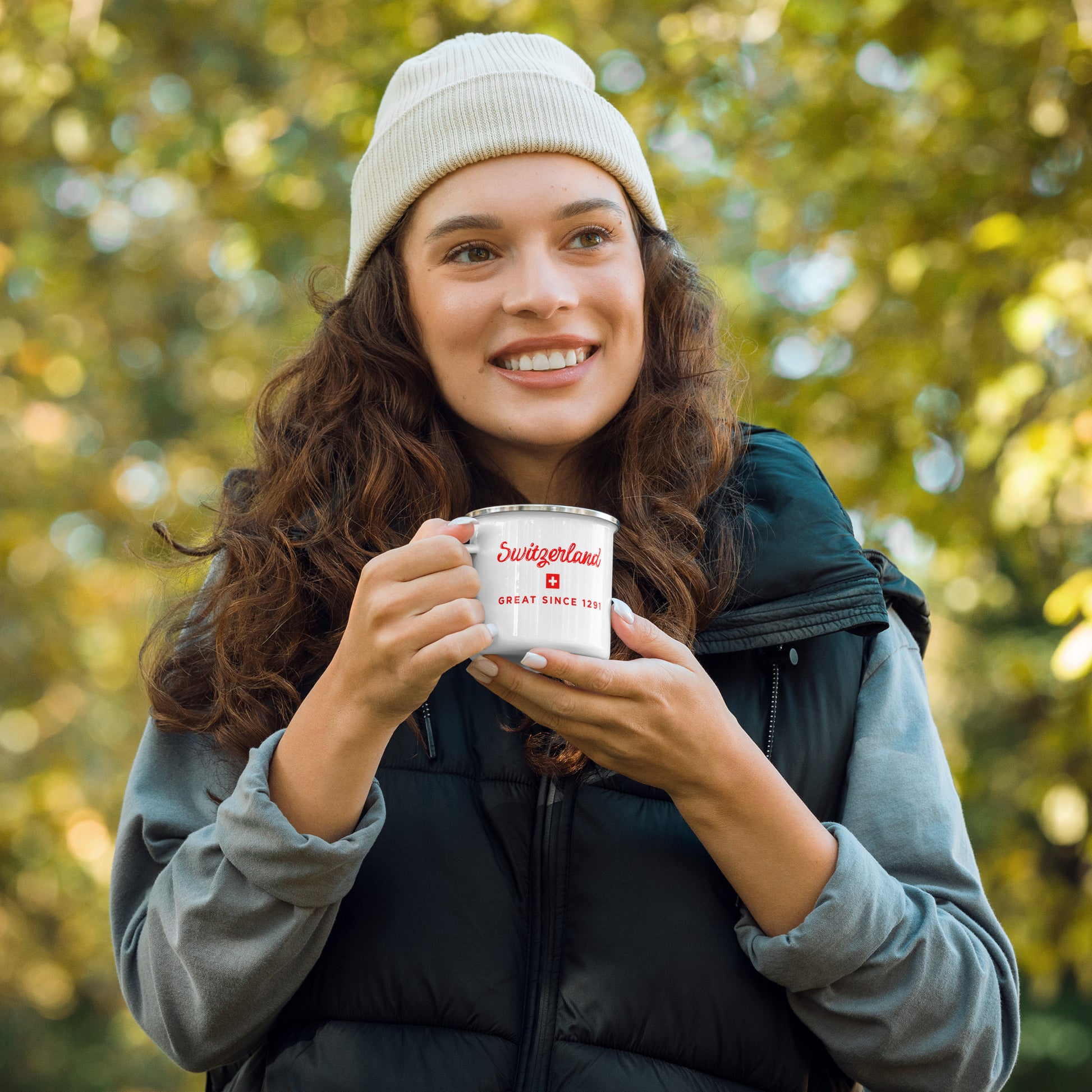 A smiling woman outdoors in a forest setting,  holding a white enamel camping mug says "Switzerland Great Since 1291".