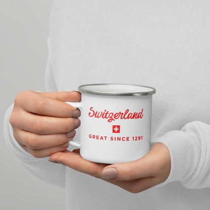 A person holding a white enamel-style mug with a silver rim. The mug is printed with the word 'Switzerland' in red script, a small Swiss flag, and the words 'GREAT SINCE 1291' in red.