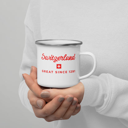 A person in a white sweatshirt holds a white enamel-style mug with a silver rim. The mug is printed with the word 'Switzerland' in red script, a small Swiss flag, and 'GREAT SINCE 1291' in red text.