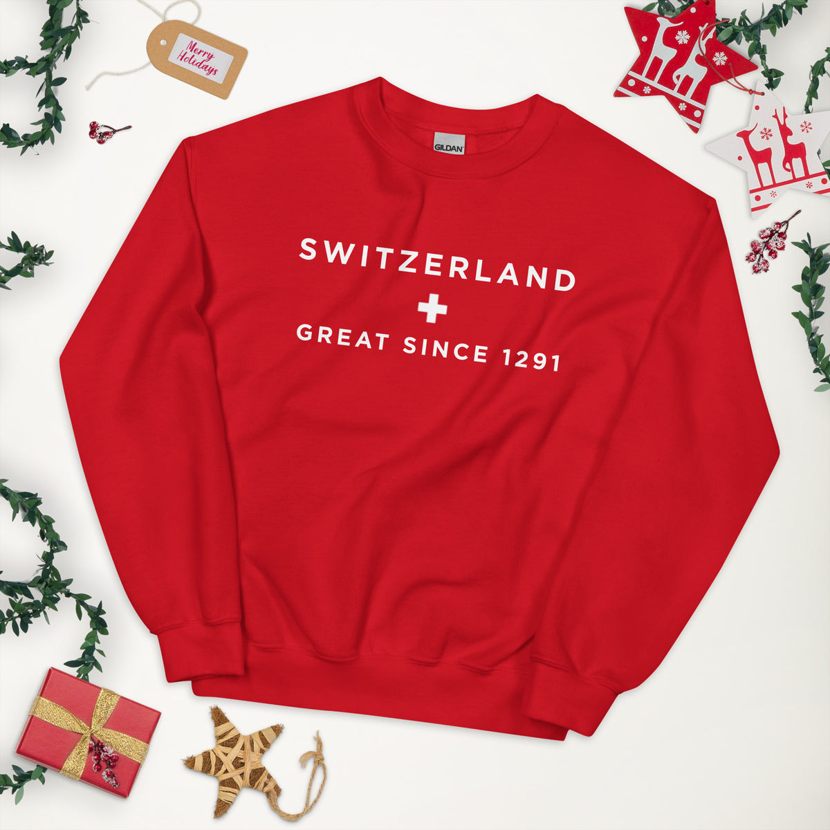 A red crewneck sweatshirt with 'SWITZERLAND + GREAT SINCE 1291' printed in white on the chest, styled in a flat lay with Christmas decorations 
