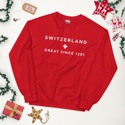 A red crewneck sweatshirt with 'SWITZERLAND + GREAT SINCE 1291' printed in white on the chest, styled in a flat lay with Christmas decorations 