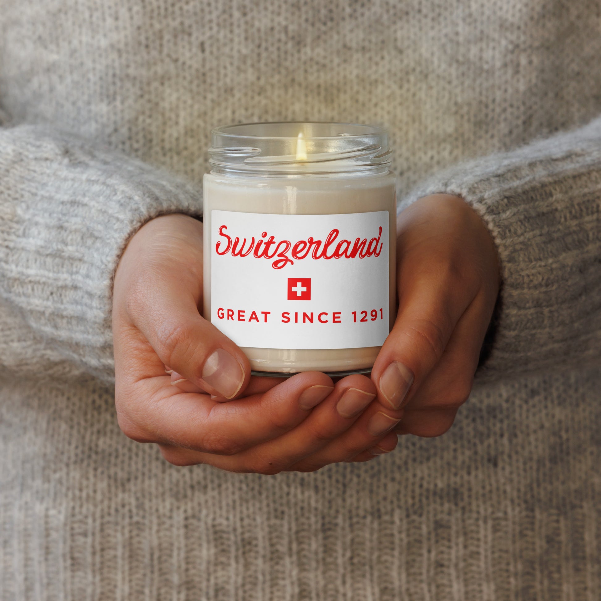 Candle in a jar labeled "Switzerland great since 1291 souvenir" held by a person wearing a textured sweater.
