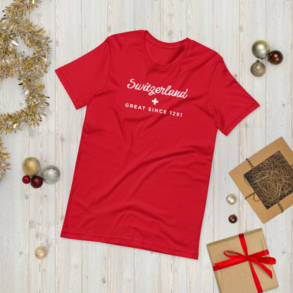 A red short-sleeve t-shirt with 'Switzerland GREAT SINCE 1291' printed in white on the chest, styled in a flat-lay on a light wood background with Christmas decorations.